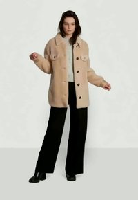 Casaco em mistura de lã bege com fecho de botão, dois bolsos no peito e um corte descontraído, usado sobre um suéter cinza claro e calças largas pretas.