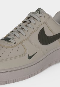 Μπεζ αθλητικό παπούτσι Nike Air Force με γκρι κορδόνια, μαύρα λογότυπα Nike swoosh στο πλάι και στη μύτη, και υφασμένο πέλμα.