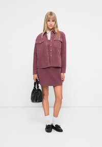 Κοντό πανωφόρι καπιτονέ και συνδυασμένη φούστα σε μωβ απόχρωση, με μπροστινές τσέπες και κουμπιά. Το μοντέλο κρατά μια μαύρη τσάντα χειρός με υφή.