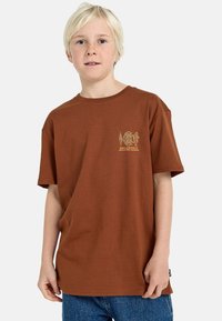 Braunes Baumwoll-T-Shirt mit kurzen Ärmeln und Rundhalsausschnitt, das auf der linken Brust ein kleines gelbes Grafikdesign zeigt. Schlichte, lockere Passform.