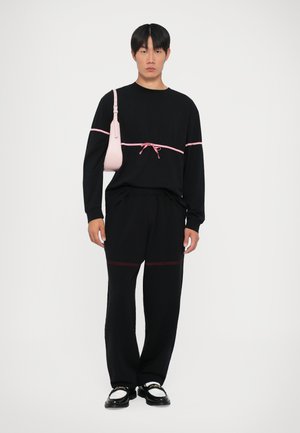 Schwarzes Langarmshirt mit pinkem Akzent und Kordelzug in der Taille, kombiniert mit schwarzen Hosen mit roter Naht. Trägt eine kleine hellrosa Tasche.