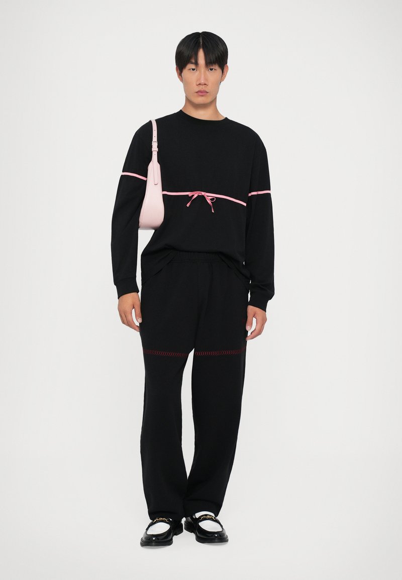 Camisa negra de manga larga con acento rosa y cintura con cordón, combinada con pantalones negros con costuras rojas. Lleva un pequeño bolso rosa claro.
