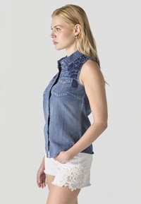 Mujer con cabello largo y rubio que lleva una camisa sin mangas de mezclilla con bordados florales y pantalones cortos blancos con detalles de encaje.