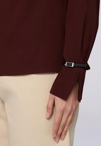 Mano appoggiata accanto a pantaloni beige, indossa una blusa a maniche lunghe bordeaux con polsini a fessura fermati da una piccola cintura nera e una fibbia d'argento.