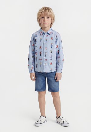 Blonder Junge steht, trägt ein hellblaues Hemd mit Cartoonfiguren, Jeansshorts und weiße Turnschuhe auf weißem Hintergrund.