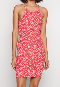 Punainen kukallinen mekko, jossa on ohuet olkaimet, korostaa vartalon muotoa ja siinä on pieniä valkoisia ja vaaleanpunaisia kukkia tekstuuripohjan päällä.