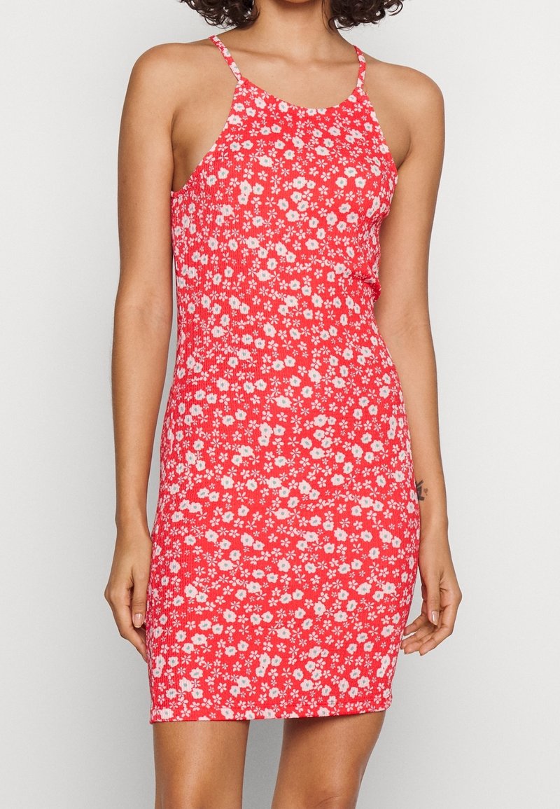 Punainen kukallinen mekko, jossa on ohuet olkaimet, korostaa vartalon muotoa ja siinä on pieniä valkoisia ja vaaleanpunaisia kukkia tekstuuripohjan päällä.