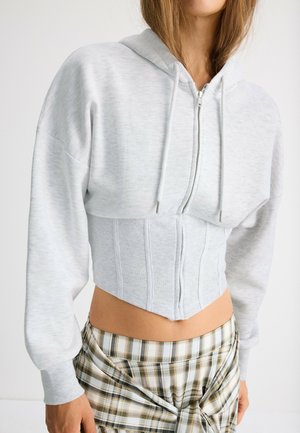 Femme portant un sweat à capuche court zippé gris clair sur un top corset côtelé, associé à un pantalon à carreaux avec une taille nouée.