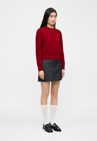 Femme debout portant un haut rouge à manches longues, une mini-jupe en denim foncé, des chaussettes blanches montantes et des mocassins noirs sur un fond blanc uni.