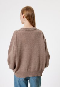 Przesadnie duży brązowy sweter z ribbowym kołnierzem i teksturowanym splotem, z obniżonymi ramionami i luźnym krojem, w zestawie z niebieskimi dżinsami.