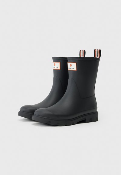 Bottes de cheville en caoutchouc noir avec semelles texturées, patch de logo orange et blanc, et deux languettes de tirage de couleurs contrastantes à l'arrière.