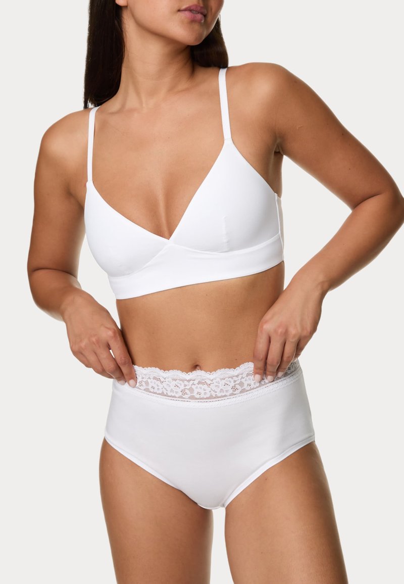 Femme portant un soutien-gorge triangle blanc et une culotte taille haute blanche avec bordure en dentelle, ajustant la ceinture de la culotte.