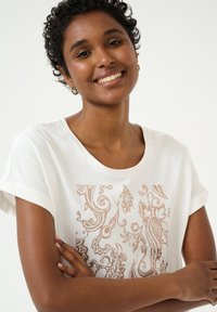 T-shirt blanc à manches courtes avec un motif paisley en rose doré sur le devant. Tissu doux et léger avec des poignets roulés.