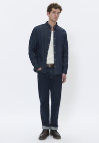 Camisa de mezclilla oscura de botones sobre una camiseta de tirantes blanca con textura, combinada con jeans de mezclilla oscuros y zapatos de cuero marrón. Un atuendo simple y casual.