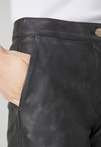 Juodos odinės kelnės su lygiu paviršiumi, turinčios šoninę kišenę ir metalinį mygtuką ant juosmens. Subtilūs spalvos pokyčiai.