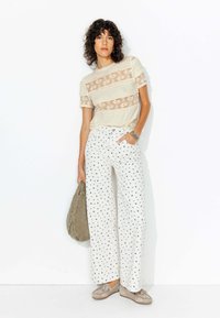 Un haut en dentelle avec des bandes horizontales crème, associé à un pantalon large blanc orné de petits motifs floraux bleus. Le mannequin tient un sac gris.