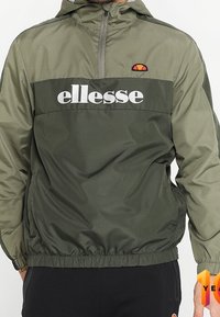 Olivově zelená větrovka s půl zipem, elastickými manžetami a lemem. Obsahuje bílé logo "ellesse" a nášivku loga na kapuci.