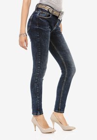 Dunkelblaue Skinny-Jeans mit strukturiertem Finish, gelben Nähten und einem mit Edelsteinen besetzten Gürtel. Kombiniert mit hellbeigen High Heels.