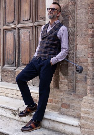 Brown plaid vest over a striped shirt, navy trousers, and sneakers. Sunglasses complete the look against a textured stone wall and wooden door.