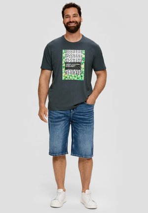 Graues T-Shirt mit grünem Grafikdruck und schwarzem Text, kombiniert mit blauen Denim-Shorts. Weiße Sneakers vervollständigen das Outfit.