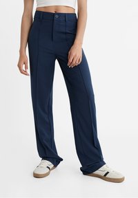 Pantalons larges navy en tissu lisse, avec une fermeture à bouton et un pli avant. Assortis à des baskets blanches avec des semelles marron.