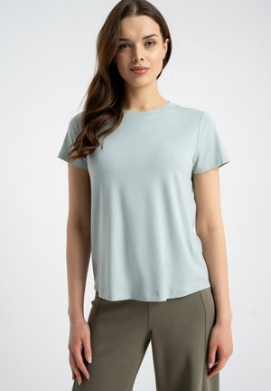 Młoda kobieta z długimi brązowymi włosami, ubrana w jasnoniebieską koszulkę z krótkim rękawem i oliwkowozielone spodnie, stojąca na tle jednolitego białego tła.