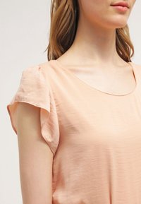 Blusa de color melocotón con mangas cortas y acampanadas y un escote amplio. La tela parece ligera y suave, con un sutil brillo.