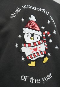 Melna auduma ar izšūtu pingvīnu, kurš valkā sarkanu mirdzošu cepuri, svītrotu šalli un dzeltenas kājas, ko ieskauj sniegpārslu motīvi un teksts.