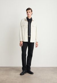 Lys beige knappet jakke med to brystlommer, kombinert med en svart skjorte og bukser. Sorte sko fullfører antrekket.