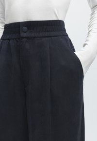 Σκούρο μπλε παντελόνι με ψηλή μέση, ελαστική μέση και κουμπί, φορεμένο με λευκό μακρυμάνικο μπλουζάκι, χέρι στην δεξιά τσέπη.