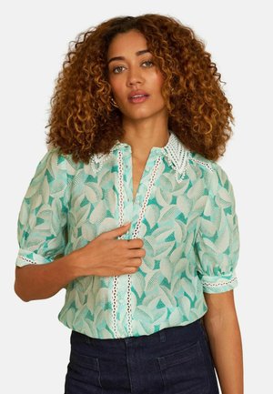 Femeie cu păr creț, purtând o bluză verde mentă cu modele și dantelă la margine și pantaloni bleumarin, pozând cu o mână aproape de guler.