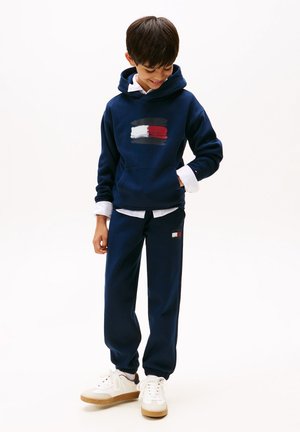 Conjunto de sudadera y pantalones de chándal azul marino con un diseño de pinceladas en blanco y rojo. Camiseta blanca debajo, combinada con zapatillas blancas con suelas beige.