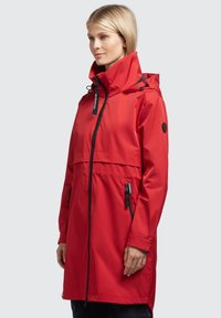 Chaqueta impermeable roja con capucha, cierre delantero con cremallera y bolsillos laterales. Presenta acentos negros y cordones ajustables. Textura exterior suave.