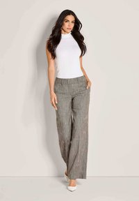 Vrouw met lang, donker haar, gekleed in een mouwloze witte coltrui, grijze wijde broek en witte puntige hakken, staande tegen een beige muur.