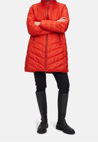 Manteau matelassé rouge avec un col haut, une fermeture éclair à l'avant, et deux poches latérales. Associé à un pantalon noir ajusté et des bottes noires montantes.