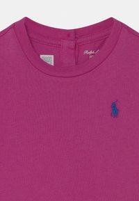 Fuchsiové bavlněné tričko s kulatým výstřihem, na levé části hrudi s modrým vyšitým logem a štítkem poblíž výstřihu.