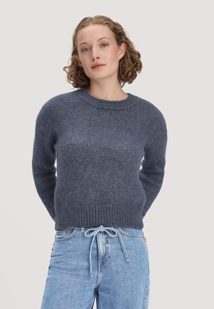 Vrouw met kort krullend haar, gekleed in een blauwe gebreide trui en lichtblauwe spijkerbroek, staand met handen achter haar rug tegen een effen achtergrond.