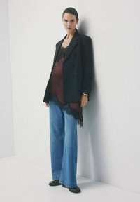 Kobieta o ciemnych włosach w czarnym blezerze, burgundowej bluzce wykończonej koronką, szerokich niebieskich dżinsach i czarnych butach oparta o białą ścianę.