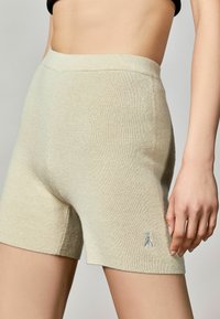 Světle béžové žebrované pletené šortky s jemnou strukturou, s logem v kontrastní barvě poblíž lemu. Středně vysoký pas.