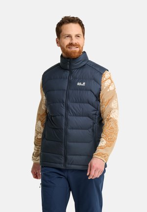Mann trägt eine marineblaue gesteppte ärmellose Weste über einem beige gemusterten Langarmhemd, steht und schaut leicht zur Seite.
