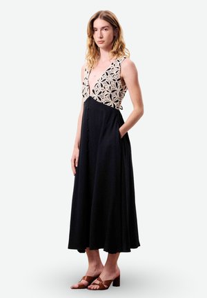 Vrouw met golvend haar die een mouwloze zwarte jurk draagt met een crème patroon en een diepe V-hals, staand met één hand in de zak en bruine sandalen met hak.