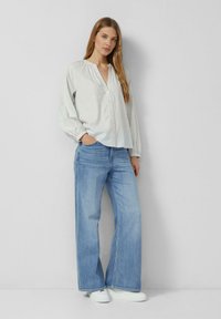 Jasnoszara koszula na guziki z marszczonym dekoltem, zestawiona z jasnoniebieskimi, szerokimi spodniami jeansowymi i białymi trampkami, na jednolitym tle.