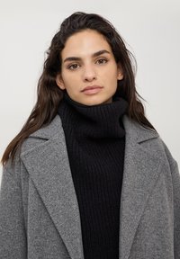 Cappotto grigio in misto lana con una silhouette strutturata, ampi risvolti, indossato sopra un maglione a collo alto nero a coste spesso.