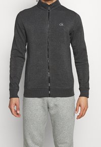 Šedá mikina na zip s vysokým límcem, žebrovanými manžetami a lemem, se malým bílým logem na levé hrudi. Měkká, texturovaná tkanina.