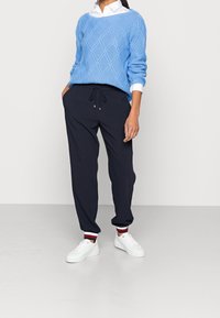 Vaaleansininen neulepusero, jossa on timanttikuvio, yhdistettynä tummansinisiin jogger-housuihin ja valkoisiin tennareihin, joissa on punaisia ja sinisiä raitoja.