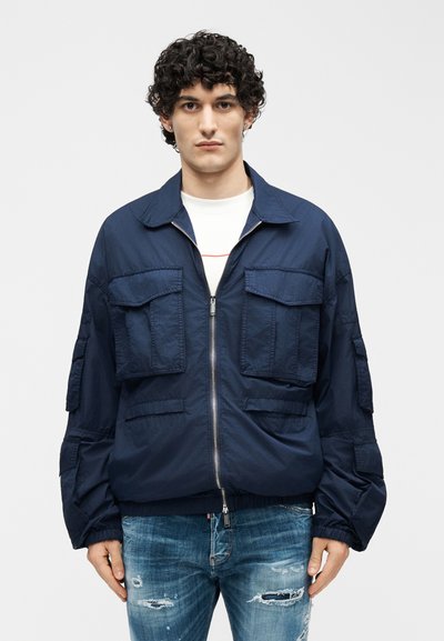 Άνδρας με σγουρά μαλλιά που φοράει μπλε σακάκι με φερμουάρ και πολλά μπροστινά τσέπες πάνω από λευκό πουκάμισο και σκισμένο μπλε τζιν.