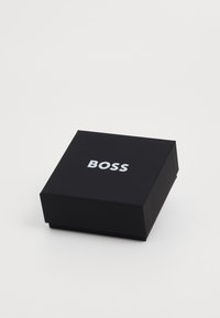 Juodas stačiakampis dėžė su matinio paviršiaus apdaila, viršuje esantis žodis "BOSS" ryškiomis sidabrinėmis raidėmis. Minimalistinis dizainas, be papildomų detalių.