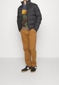 Person klädd i en svart puffjacka, färgglad mönstrad tröja, senapsgula byxor och svarta sneakers med vita detaljer som står mot en enkel bakgrund.