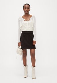 Un cardigan en tricot blanc sur un haut crème à motifs, associé à une jupe en velours côtelé marron. Les accessoires comprennent un petit sac beige et des bottes de cowboy blanches.
