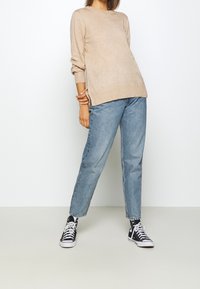 Pull en tricot beige à manches longues, col rond, fentes latérales ; associé à un jean droit bleu clair et des baskets montantes noires.